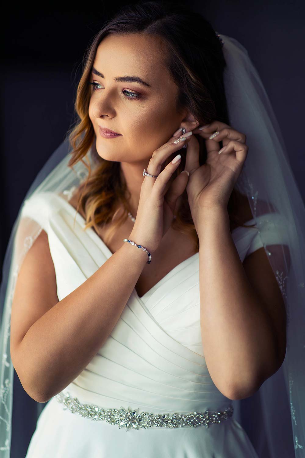 Bride gently putting in her earrings with her veil over her shoulders
