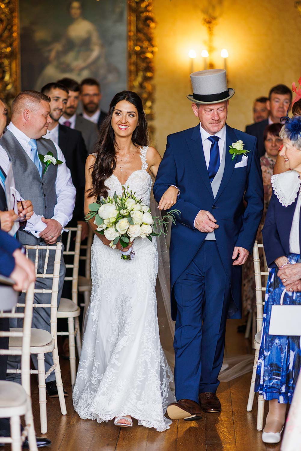 Smiling bride walks down the aisle with her dad who is wearing a grey top hat.