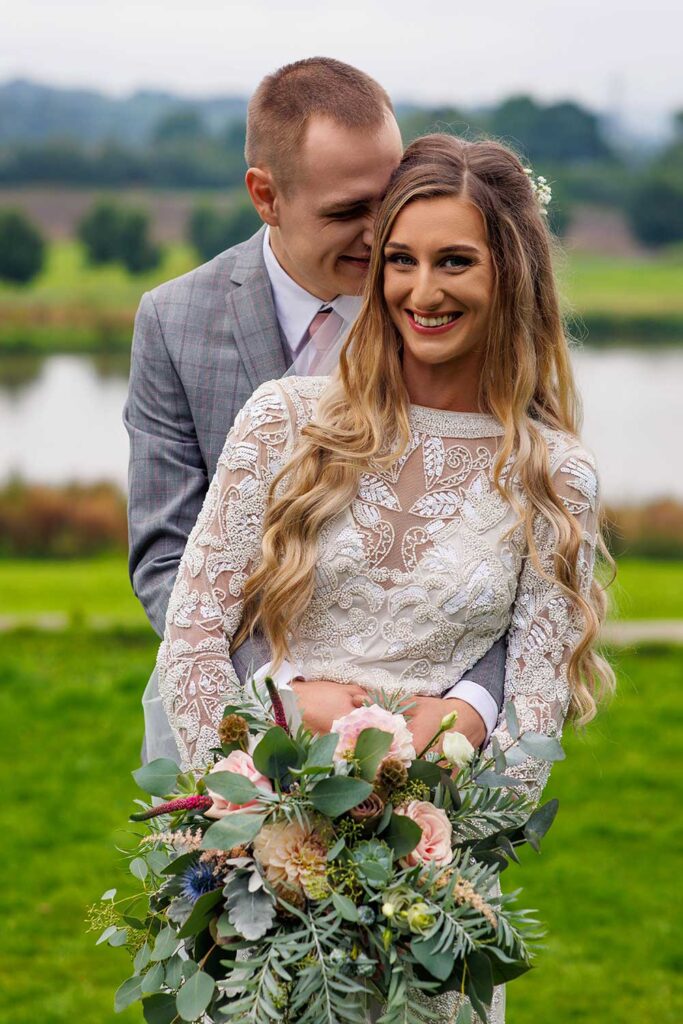 Beautiful bride smiling holding bouquet whilst groom whispers something funny into her ear