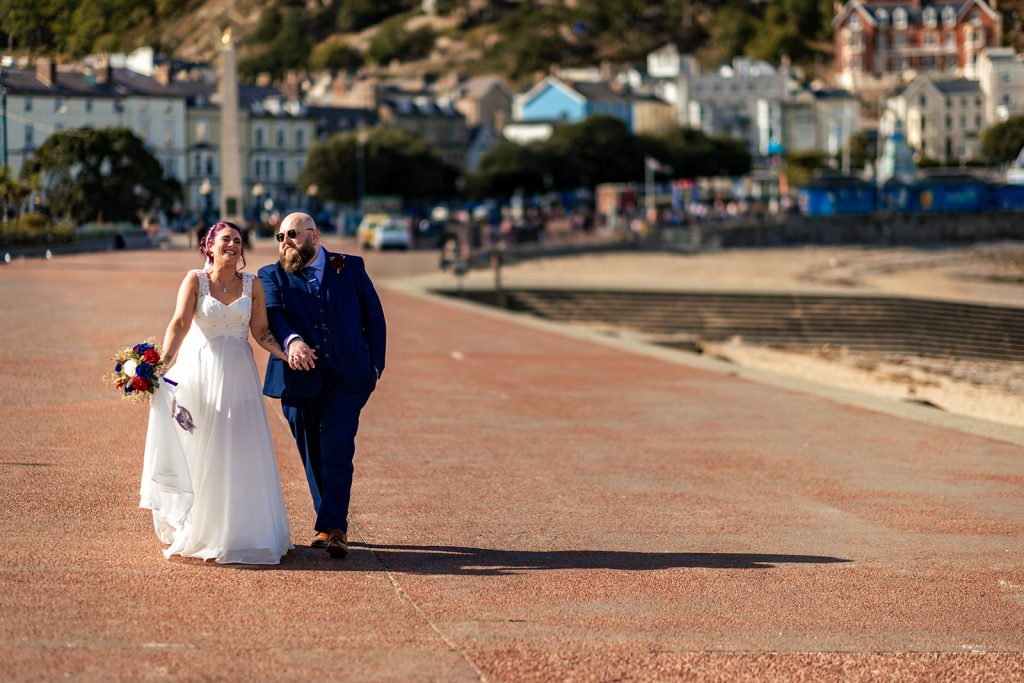 Bride and groom walking and laughing as they walk down llandudno promenade