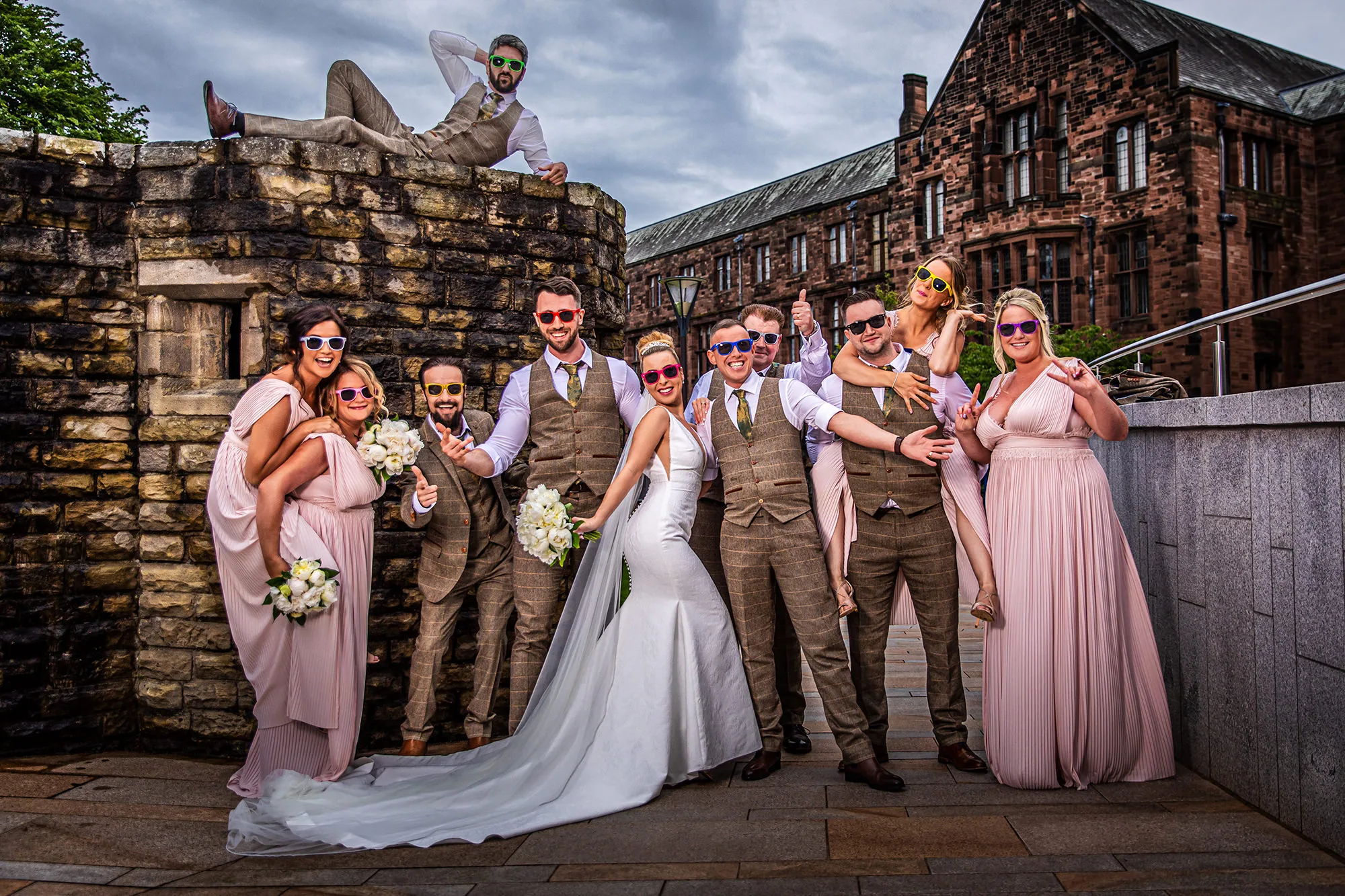 Bride and wedding party in colourful sunglasses celebrating beside a stone building