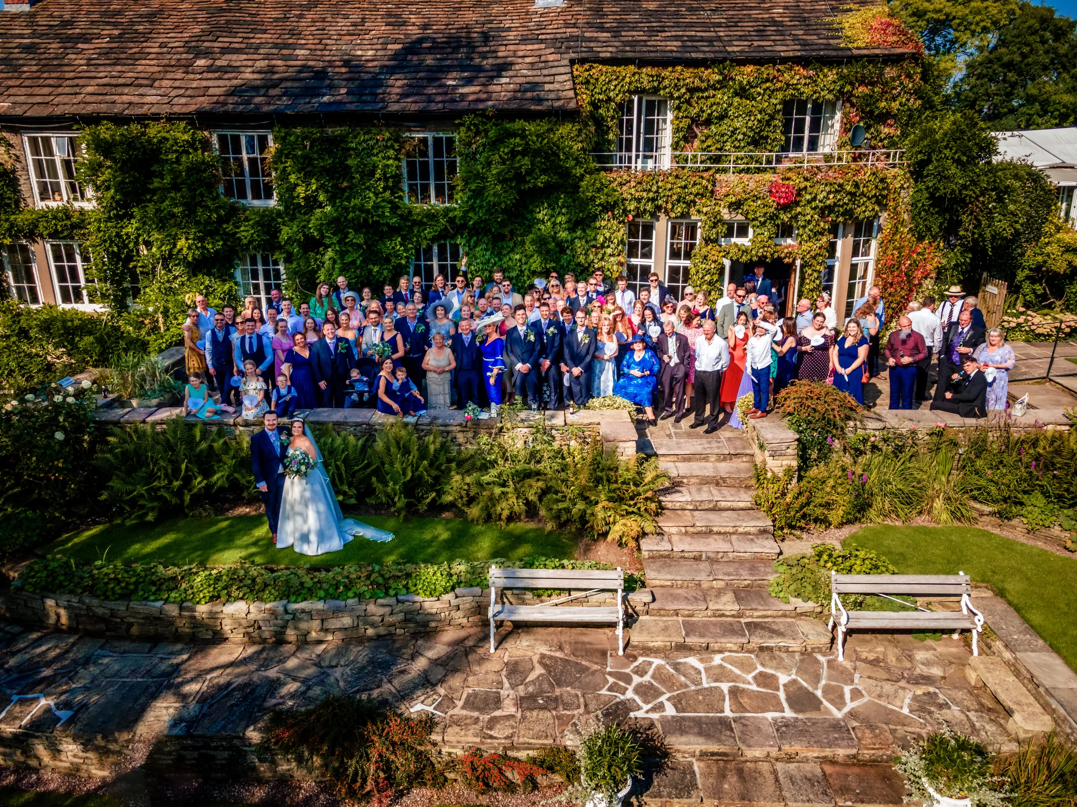 Wedding couple and guests gathered outside an ivy-covered country house, photographed from above