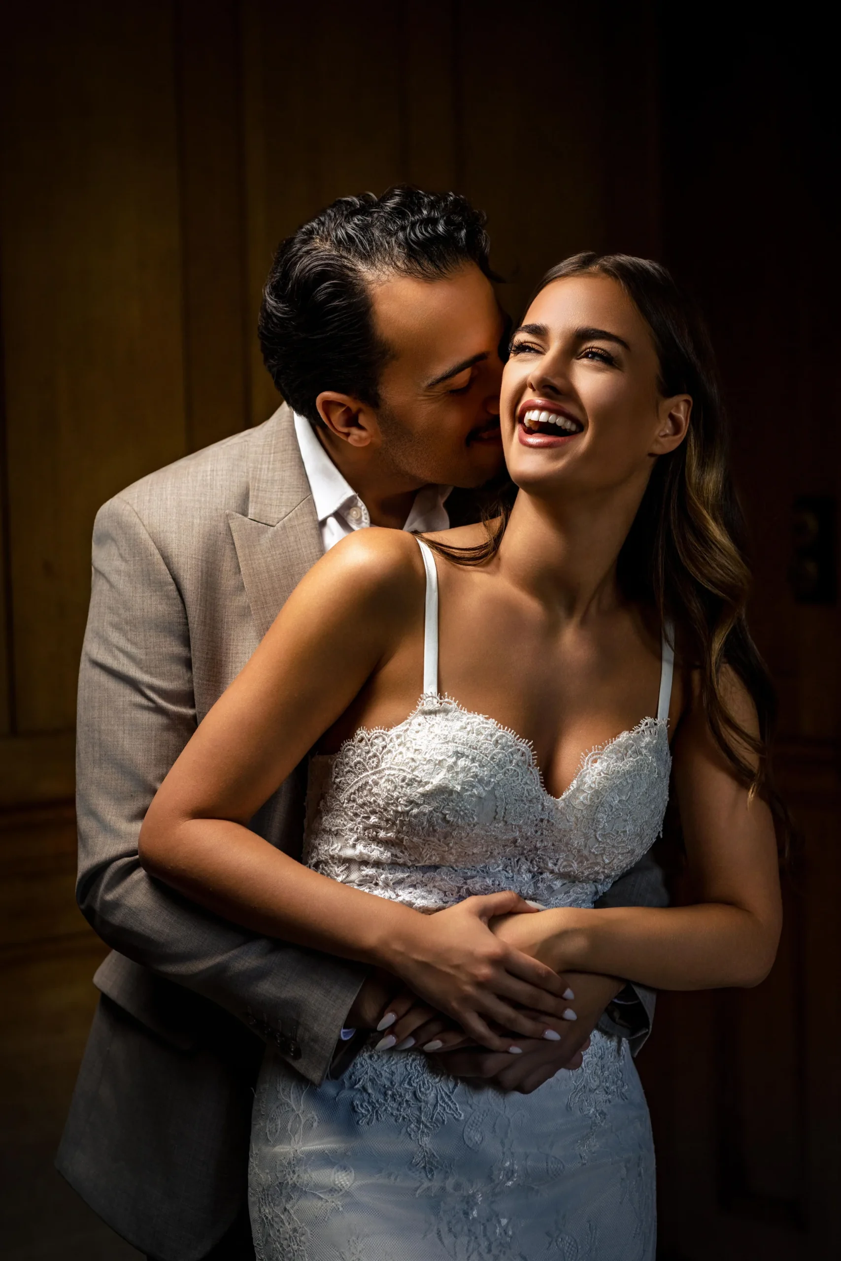 Groom hugs the laughing bride from behind in a warm indoor portrait