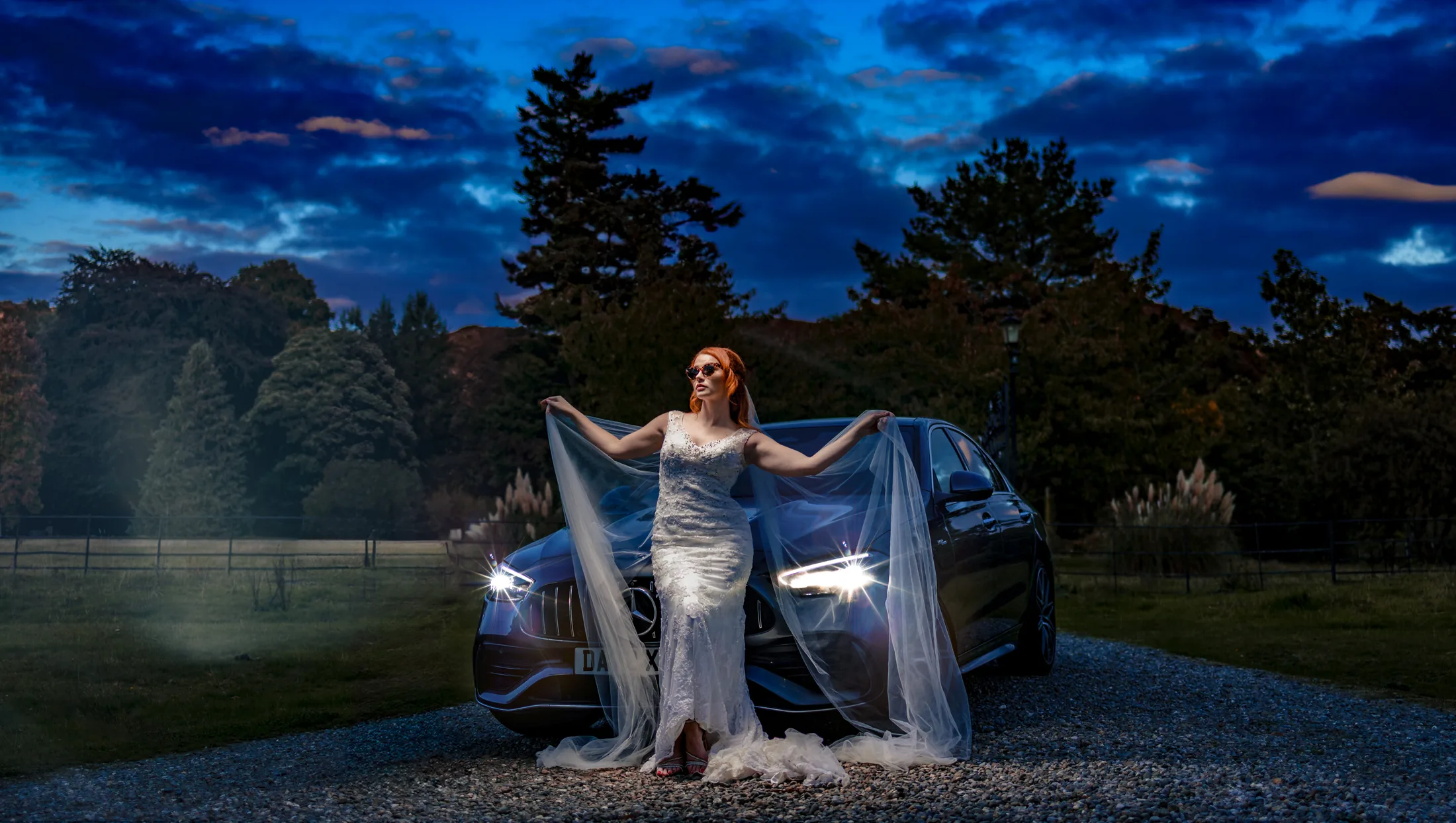 Bride holding her veil beside a car at a North West wedding, under a blue evening sky