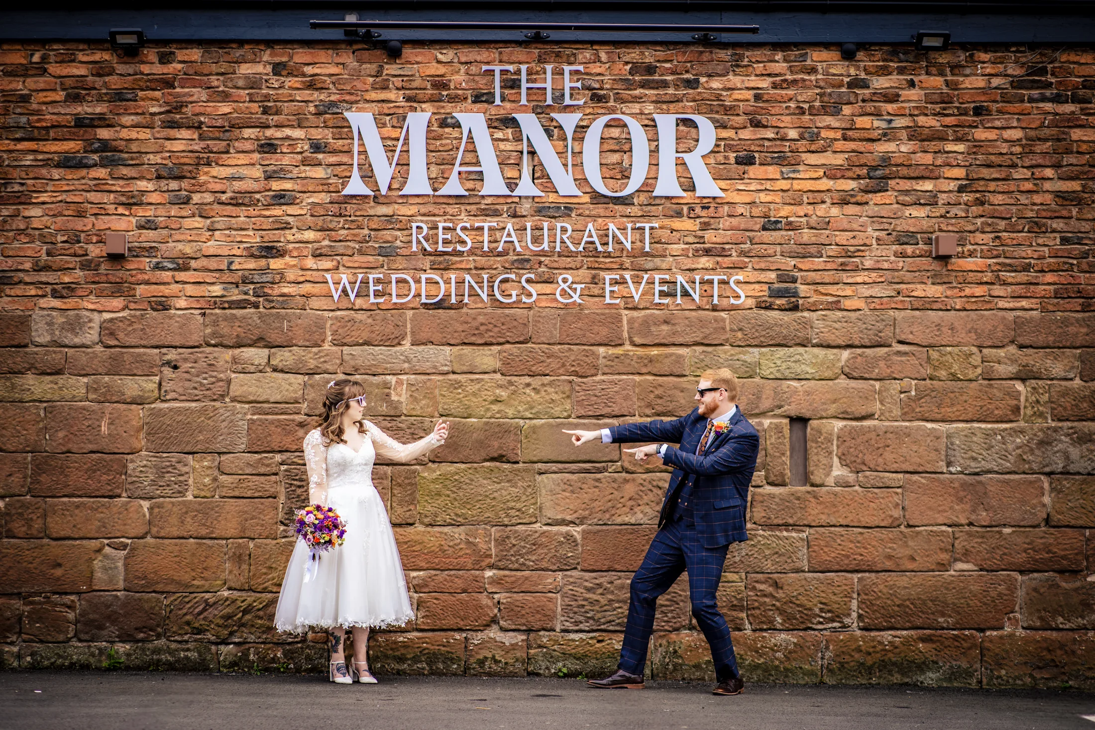 Bride and groom playfully pointing at each other beneath The Manor wedding venue sign