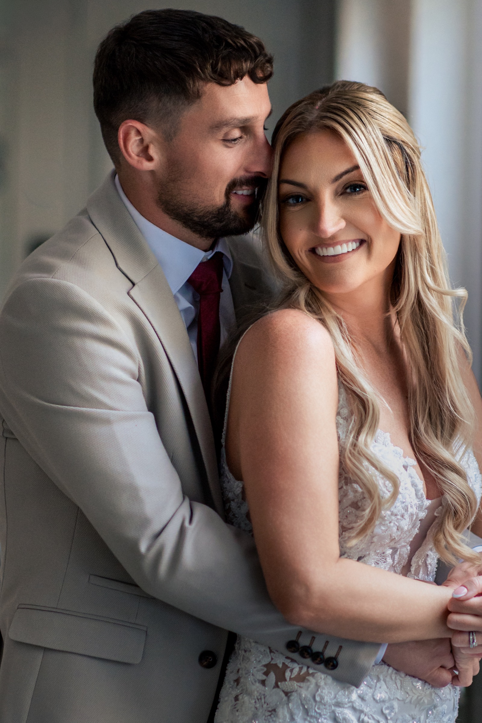 Bride smiling as the groom embraces her in soft window light